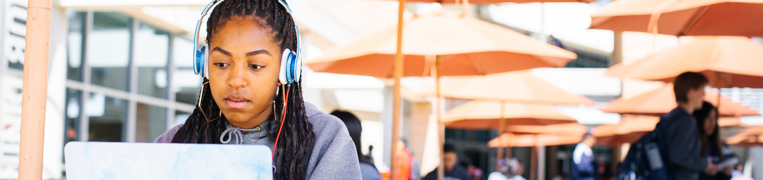 A female student wearing over ear headphones sits at a table while looking at her laptop screen