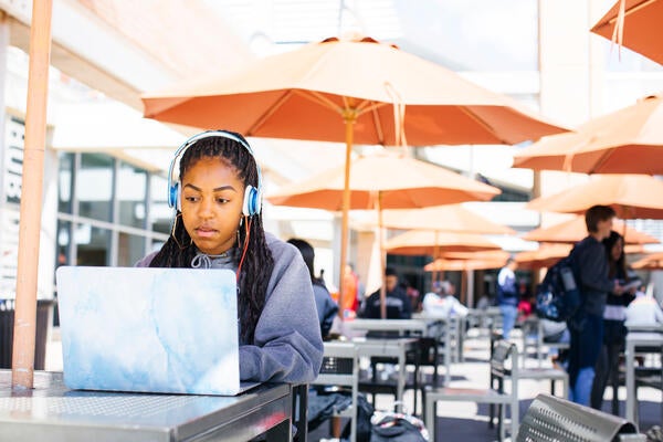 A female student wearing over ear headphones sits at a table while looking at her laptop screen